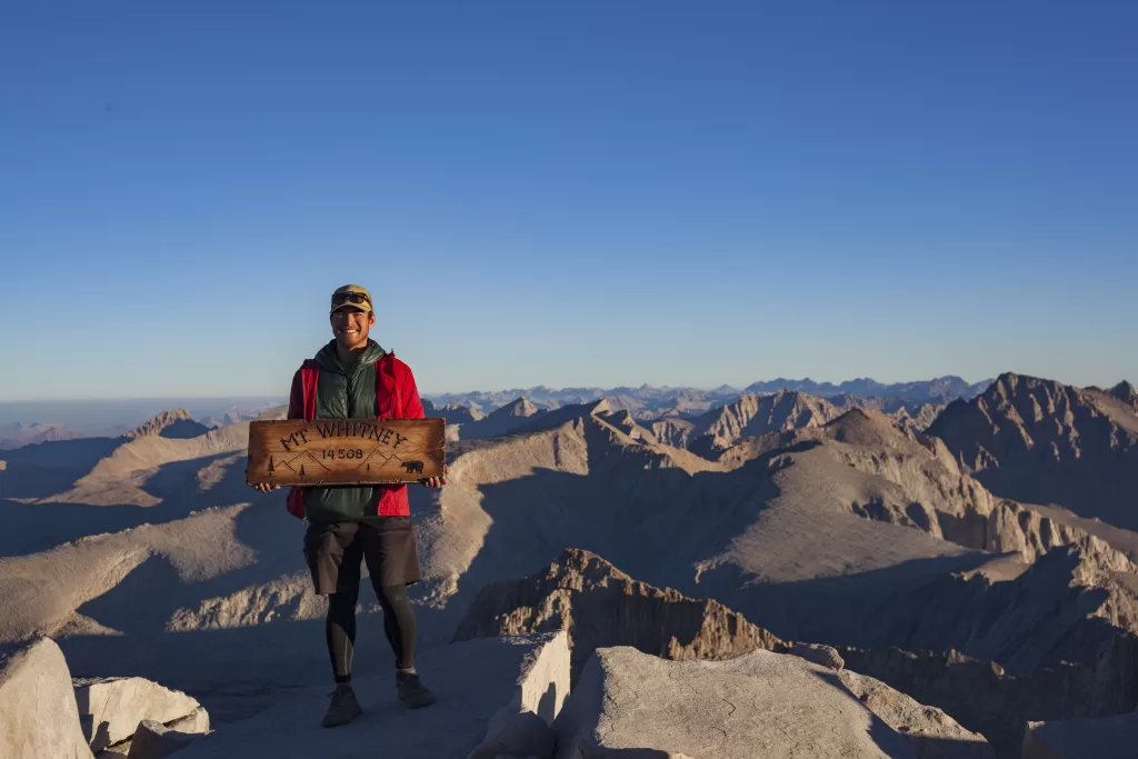 Andrew smiling wide and standing on the top of Mt. Whitney holding a Mt. Whitney sign having just completed the John Muir Trail. 