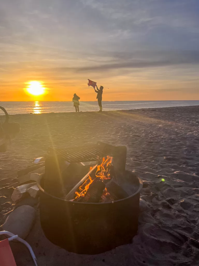 A campfire buring and two people flying a kite as the sun sets over the Pacific Ocean during our Point Mugu State Park camping trip. 