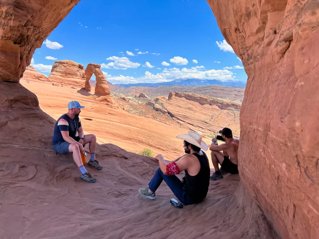 Three hikers sitting in the shade while on an adventure, viewing Delicate Arch in Arches National Park