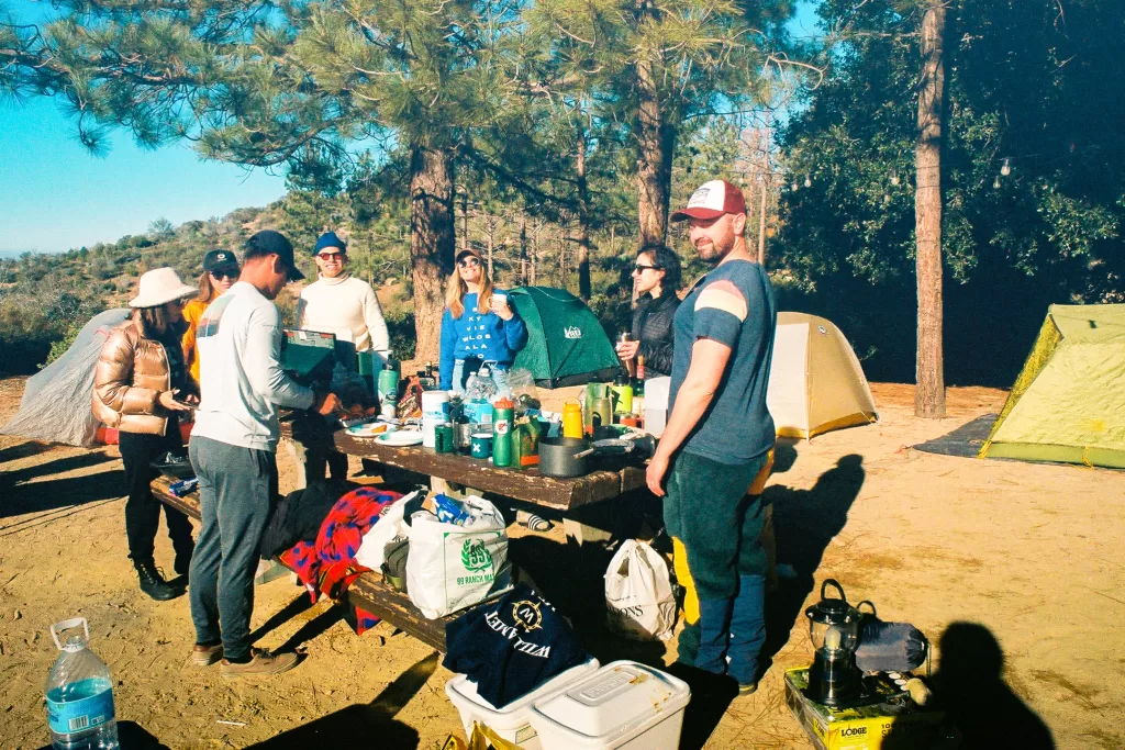 A group of friend standing around a picnic table while camping. 