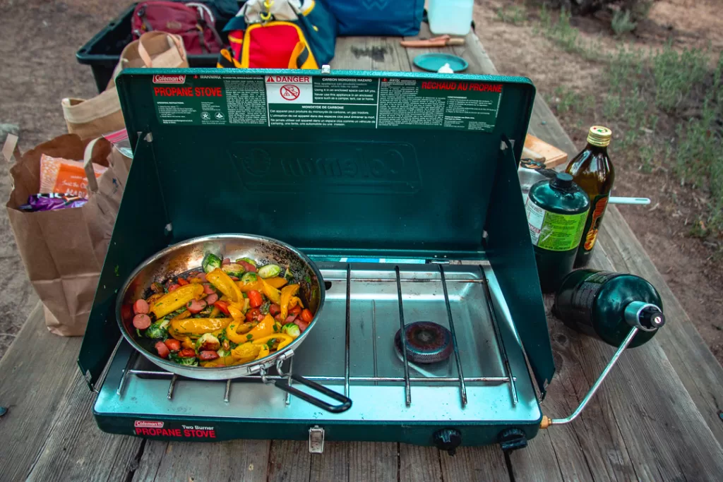 A Coleman stove with a pan and veggies sautéing. 