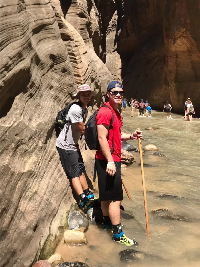 Two hikers wearing canyoneering boots, neoprene socks and a walking stick as they hike the Narrows.